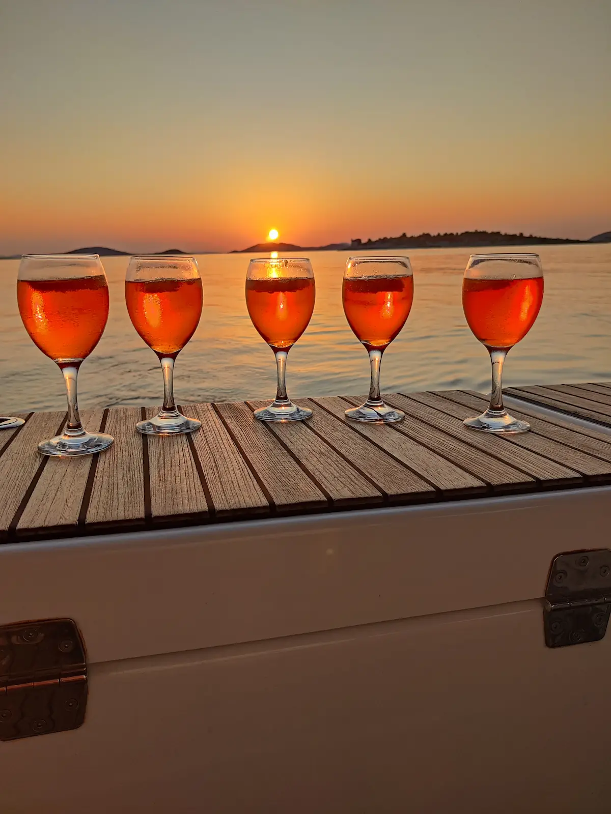 Fünf Aperol-Gläser auf Teakdeck, Sonne berührt den Horizont zwischen den Inseln