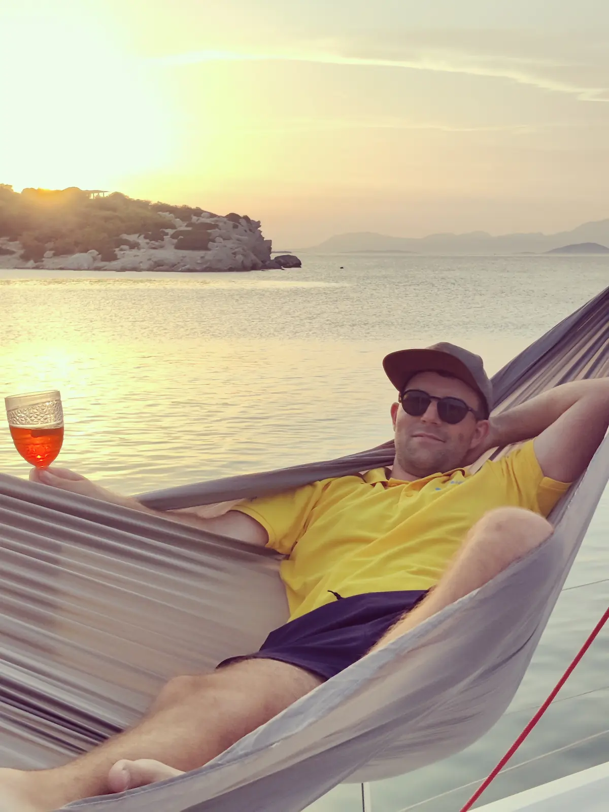 Moritz liegt mit einem Aperol in der Hängematte auf einer Segelyacht beim Sonnenuntergang in Griechenland