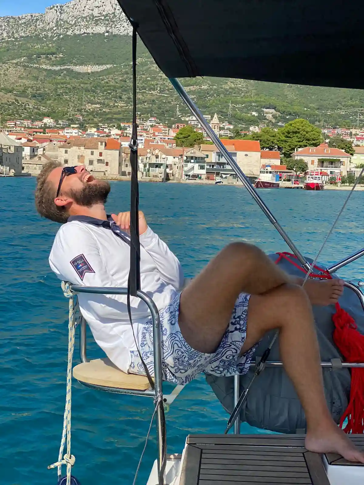 Moritz lacht entspannt auf dem Bug einer Segelyacht vor einem kroatischen Hafenort