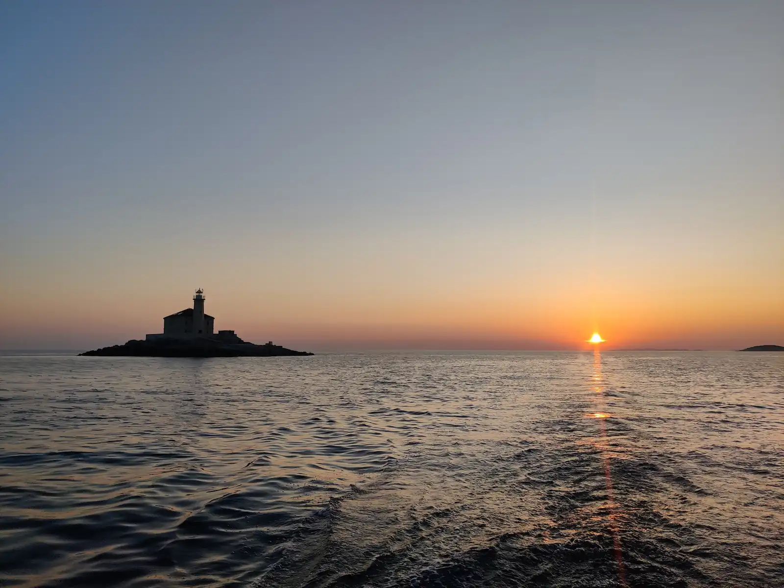 Leuchtturm als Silhouette links, Sonne sinkt rechts ins Meer, Kielwasser im Vordergrund