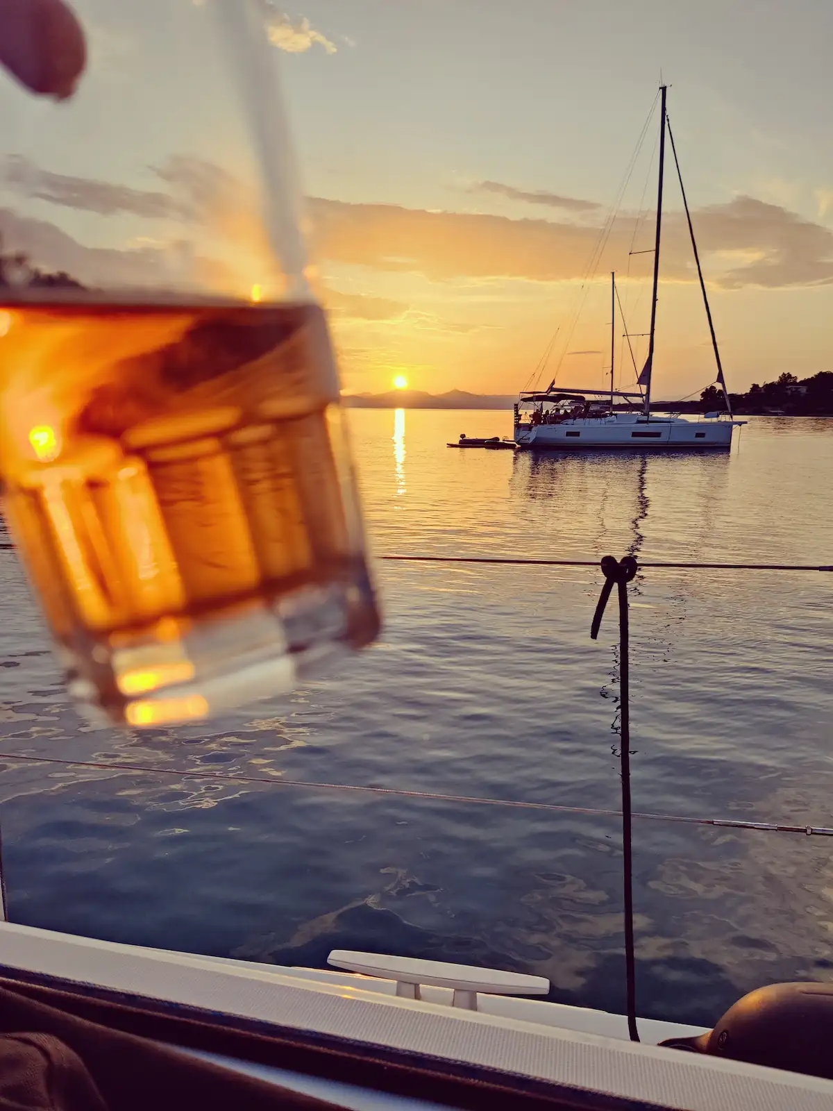 Aperol-Glas im Vordergrund, Segelyacht ankert im Sonnenuntergang auf spiegelndem Wasser